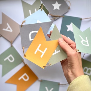 Puede incluir: Una pancarta con banderas que deletrean "Happy" en letras blancas sobre un fondo amarillo, gris y azul.