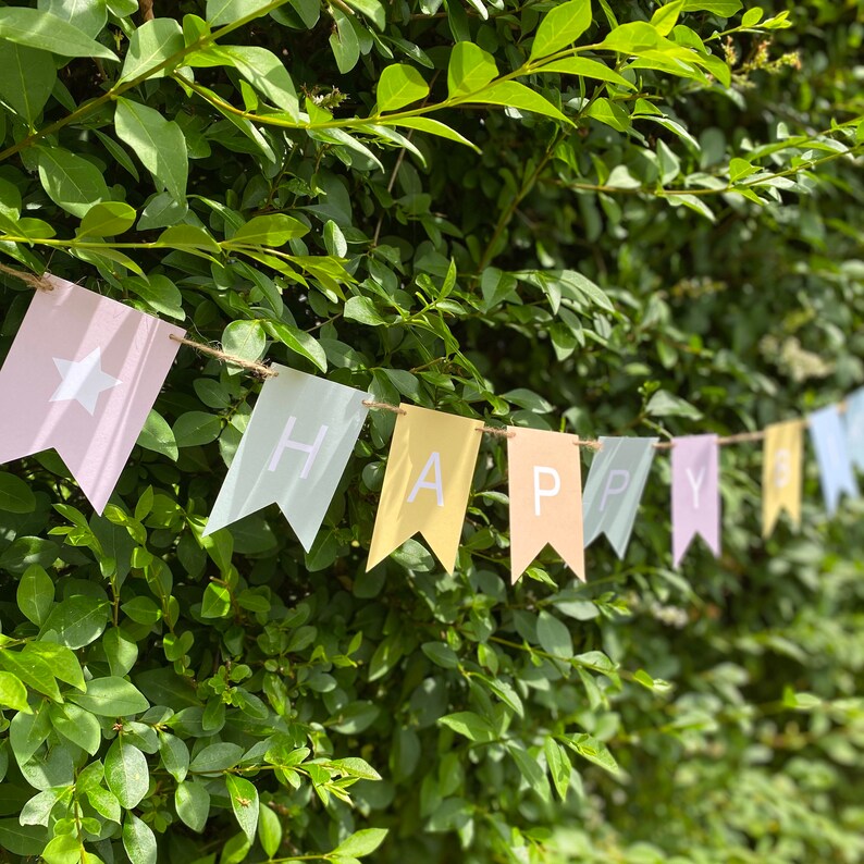 Puede incluir: Una pancarta de colores pastel con la palabra "HAPPY" escrita en ella. La pancarta est&aacute; colgada en un arbusto verde.