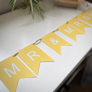 Puede incluir: Una pancarta amarilla con letras blancas que deletrean "MR & MRS" colgada en una superficie blanca.