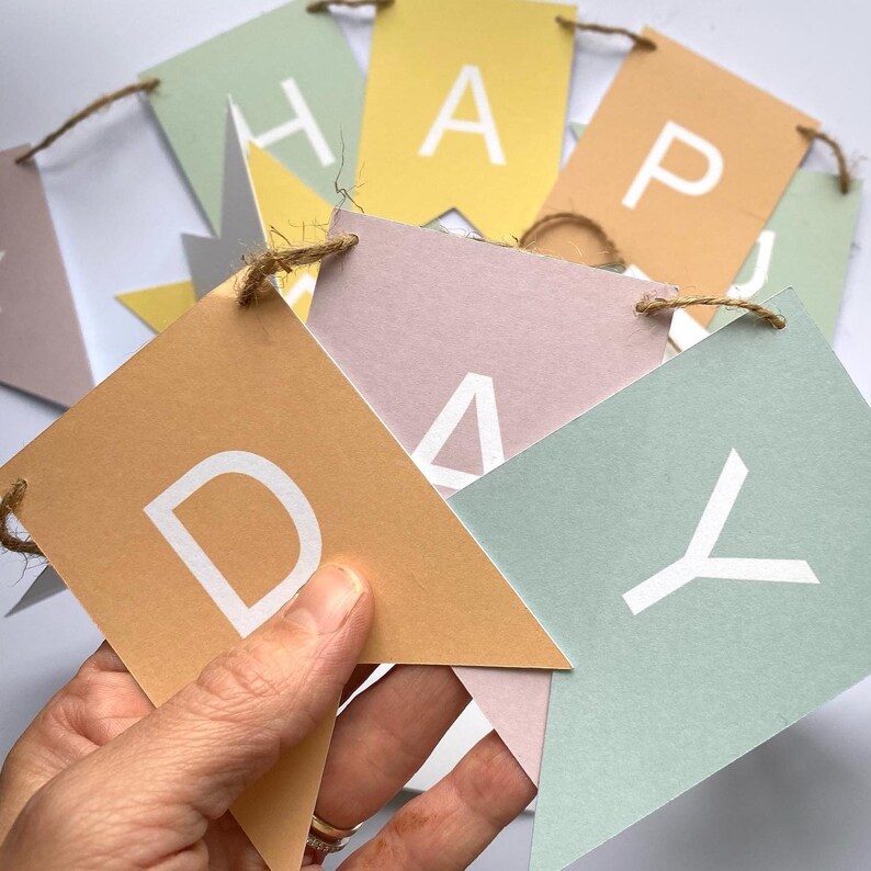 Puede incluir: Una pancarta de papel de color pastel con la palabra "Happy Day" escrita en letras blancas. La pancarta est&aacute; enhebrada con un cordel.