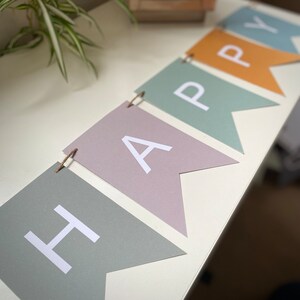 Op de afbeelding: Een pastelkleurig banner met het woord "HAPPY" gespeld in witte letters op elke vlag. De vlaggen zijn aan elkaar geregen met touw.