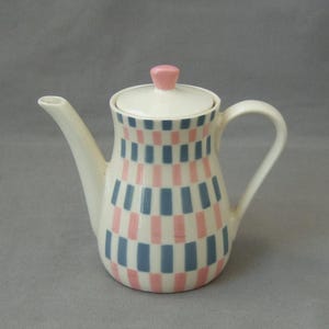 Op de afbeelding: Een witte theepot van keramiek met een roze deksel en een geometrisch patroon van roze en blauwe strepen.