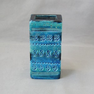 Pode incluir: Um vaso retangular de cerâmica turquesa com uma abertura quadrada. O vaso apresenta uma superfície texturizada com padrões geométricos repetidos em tons de azul e verde-azulado. A borda superior é cinza escuro. Cerca de 15 cm de altura.