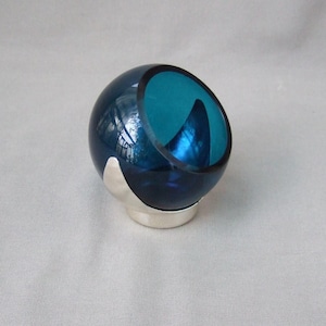 Può includere: Una piccola ciotola di vetro blu turchese con una base argentata. La ciotola ha la forma di una sfera con una sezione tagliata per creare una forma di ciotola.
