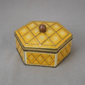 Puede incluir: Una caja de hojalata hexagonal amarilla con una tapa estampada y un pomo de madera. La tapa tiene un patrón de diamantes con detalles dorados.
