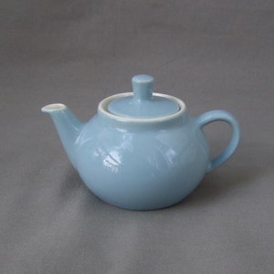 Può includere: Una teiera in ceramica blu chiaro con un coperchio bianco. La teiera ha un corpo arrotondato e un beccuccio laterale.