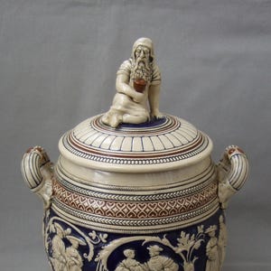 以下が含まれることがあります： クリーム、青、茶色の陶器製の蓋付きジャー。 蓋には座っている人物と装飾的なバンドがあります。 ジャーの本体はレリーフシーンとハンドルで飾られています。