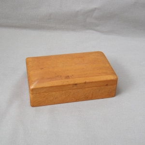 Peut inclure: Petite boîte rectangulaire en bois clair avec couvercle à charnière. Le bois a une finition lisse et polie. La boîte est vide et posée sur une surface unie de couleur claire.