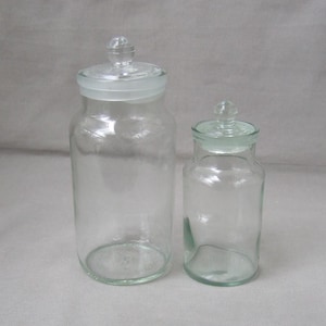 May include: Two clear glass apothecary jars with glass lids. The jars are different sizes, with the larger jar on the left and the smaller jar on the right. The jars are empty and sit on a white surface.
