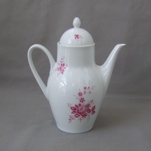 Puede incluir: Cafetera de porcelana blanca con tapa y diseño floral en tonos rosados. La cafetera tiene un cuerpo redondeado y un pico en el lateral.