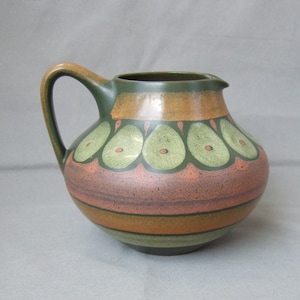 Può includere: Un'anfora in ceramica verde e marrone con un design geometrico. L'anfora ha una base larga e uno stretto beccuccio.