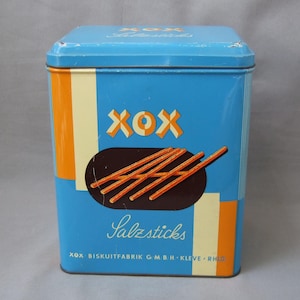 Peut inclure: Une boîte en métal bleue et orange avec le mot "XOX" et le texte "Salzsticks" imprimé dessus. La boîte contient des bâtonnets salés et porte le texte "XOX Biskuttfabrik G.m.b.H. Kleve - Rhld." imprimé au bas.