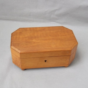 May include: A light-coloured wooden box with an octagonal lid and a keyhole. The box has a simple design and is likely used for storing small items. The box is made of wood.