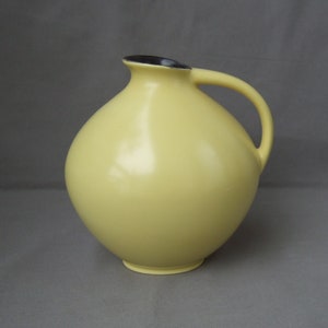 May include: A yellow ceramic vase with a black rim and a handle. The vase has a rounded, bulbous shape.