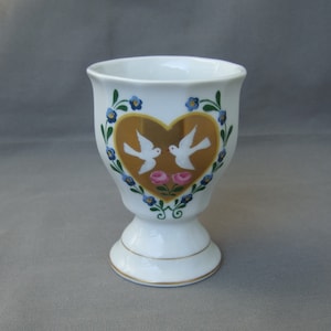 K&ouml;nnte beinhalten: Wei&szlig;e Keramiktasse mit einem goldenen Herzdesign mit zwei wei&szlig;en Tauben und rosa Rosen. Die Tasse hat einen goldenen Rand und ein blaues und gr&uuml;nes Blumenmuster.
