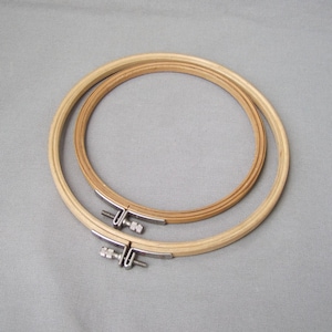 Könnte beinhalten: Zwei hölzerne Stickrahmen. Der größere Reifen ist hellbraun, der kleinere Reifen ist dunkler braun. Beide Reifen haben einen Metallverschluss zum Spannen des Stoffes. Auf grauem Hintergrund.