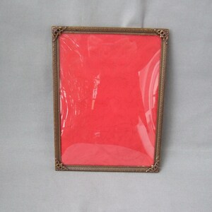 Könnte beinhalten: Rechteckiger antiker Messing-Bilderrahmen mit roter Rückwand. Der Rahmen hat dekorative Eckakzente und eine transparente Schutzhülle. Ein Vintage-Wohnaccessoire.