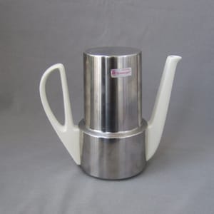 May include: A silver and white coffee pot with a stainless steel body and a white porcelain spout and handle. The pot has a label that reads "Cromargan"