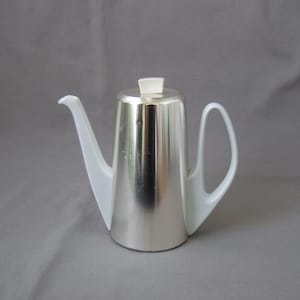 Può includere: Una teiera moderna argentata e bianca con beccuccio e manico curvi. La teiera ha un corpo lucido e metallico e un beccuccio e manico bianchi e angolari. Il coperchio è bianco e rettangolare. La teiera è probabilmente in metallo e ceramica.