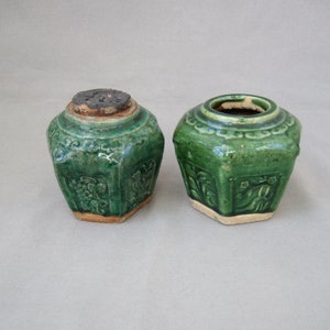 May include: Two small, green ceramic jars with intricate floral designs. The jars have a hexagonal shape and are glazed in a rich green colour.