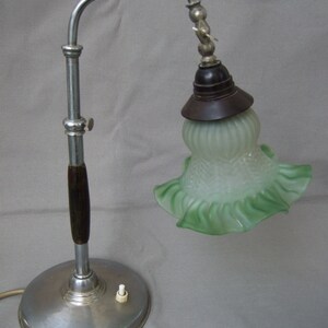 Op de afbeelding: Een tafellamp met een ronde zilverkleurige metalen voet, een centrale chromen stang met een donkerhouten accent en een verstelbare gebogen arm. De lamp heeft een donkerbruine fitting en een mat, lichtgroene glazen kap met een golvende rand.
