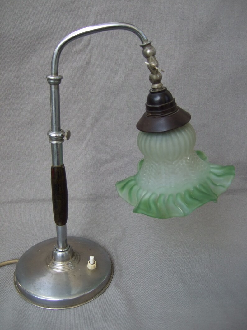 Op de afbeelding: Een tafellamp met een ronde zilverkleurige metalen voet, een centrale chromen stang met een donkerhouten accent en een verstelbare gebogen arm. De lamp heeft een donkerbruine fitting en een mat, lichtgroene glazen kap met een golvende rand.