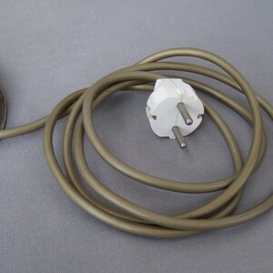 Op de afbeelding: Een metalen ronde voet verbonden met een lang, opgerold, lichtbruin elektrisch snoer. Het snoer eindigt in een witte, tweepolige stekker voor standaard stopcontacten, voorzien van twee ronde metalen pinnen en een randaardingscontact.