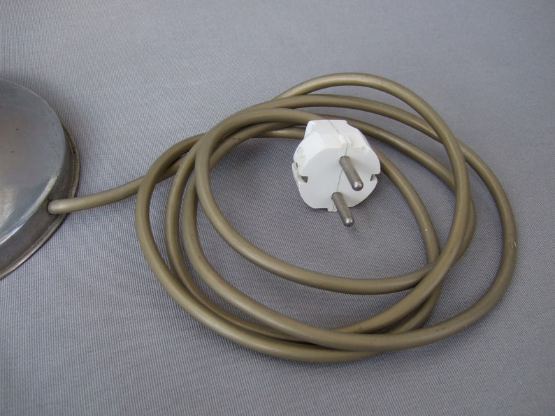 Op de afbeelding: Een metalen ronde voet verbonden met een lang, opgerold, lichtbruin elektrisch snoer. Het snoer eindigt in een witte, tweepolige stekker voor standaard stopcontacten, voorzien van twee ronde metalen pinnen en een randaardingscontact.