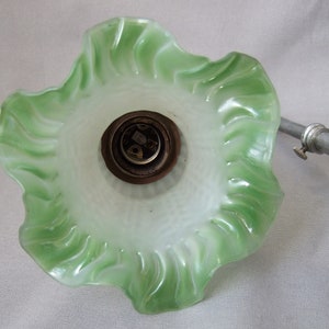 Op de afbeelding: Een golvende glazen lampenkap met een witte binnenkant die overloopt in een felgroene, geschulpte rand. De kap is bevestigd aan een metalen fitting met een donkerbruine basis en een zilverkleurige metalen arm die naar de zijkant uitsteekt.