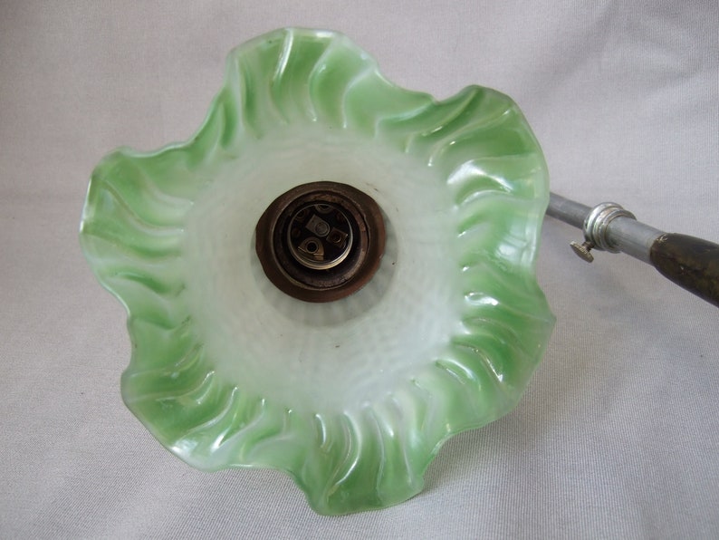 Op de afbeelding: Een golvende glazen lampenkap met een witte binnenkant die overloopt in een felgroene, geschulpte rand. De kap is bevestigd aan een metalen fitting met een donkerbruine basis en een zilverkleurige metalen arm die naar de zijkant uitsteekt.