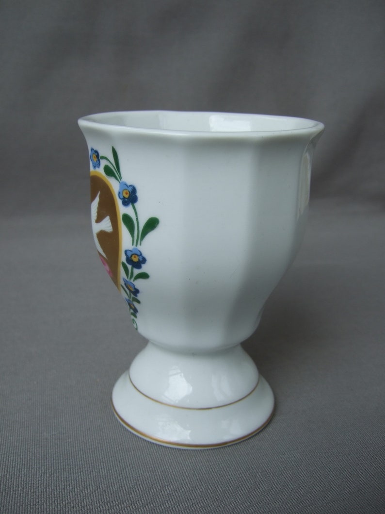 K&ouml;nnte beinhalten: Ein wei&szlig;er Porzellan-Eierbecher mit goldfarbenem Rand und einem Blumendesign mit blauen Blumen und braunen V&ouml;geln.