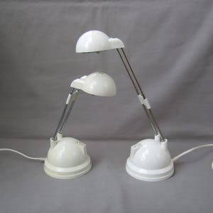 Può includere: Due lampade da scrivania bianche con bracci regolabili e paralumi a cupola. Le lampade hanno una base arrotondata e un braccio articolato color argento. Ogni lampada ha un cavo di alimentazione. Lo sfondo è grigio.