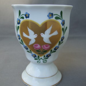 K&ouml;nnte beinhalten: Ein wei&szlig;er Porzellan-Eierbecher mit einem goldenen Herzdesign, das zwei wei&szlig;e Tauben und rosa Rosen zeigt. Das Herz ist von einem blauen und gr&uuml;nen floralen Rand umgeben.