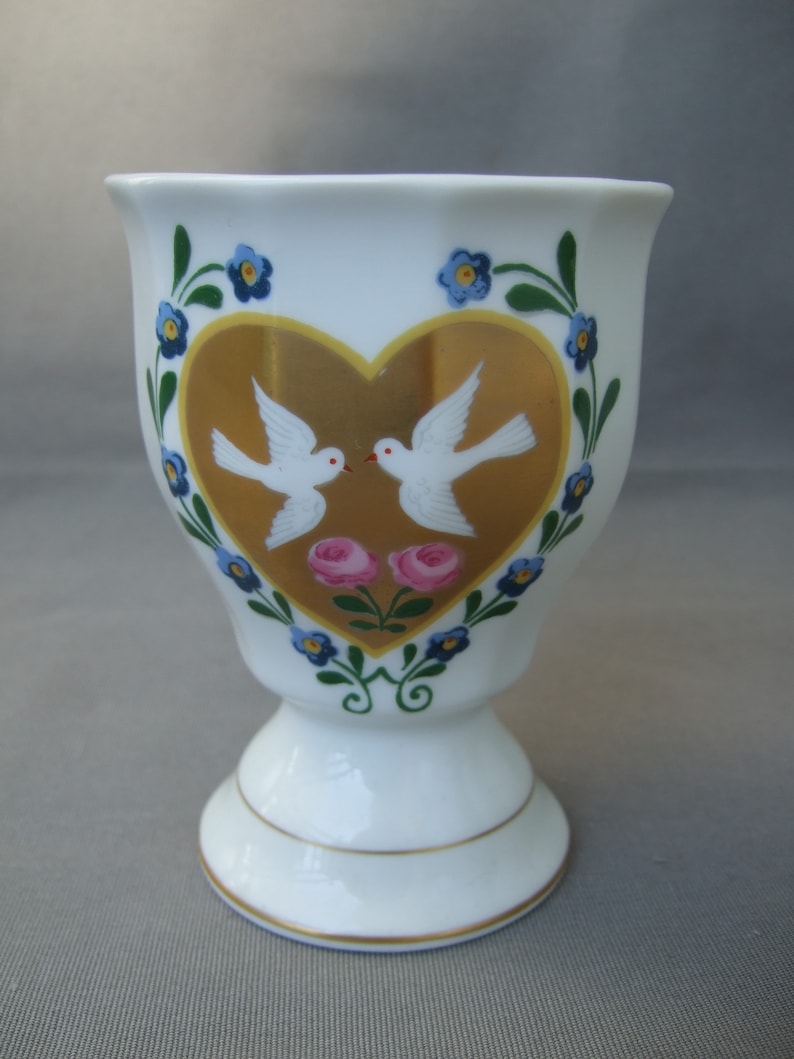 K&ouml;nnte beinhalten: Ein wei&szlig;er Porzellan-Eierbecher mit einem goldenen Herzdesign, das zwei wei&szlig;e Tauben und rosa Rosen zeigt. Das Herz ist von einem blauen und gr&uuml;nen floralen Rand umgeben.