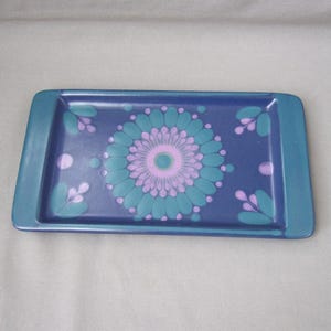 Puede incluir: Bandeja rectangular de cerámica con borde azul y diseño floral en el centro. El diseño floral presenta una flor turquesa y rosa con hojas turquesas y puntos rosas sobre fondo azul oscuro.