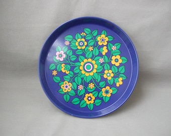 May include: A round, deep blue serving tray featuring a central, symmetrical floral pattern. The design consists of stylized yellow and pink flowers with green leaves arranged in a circular motif across the surface of the tray.