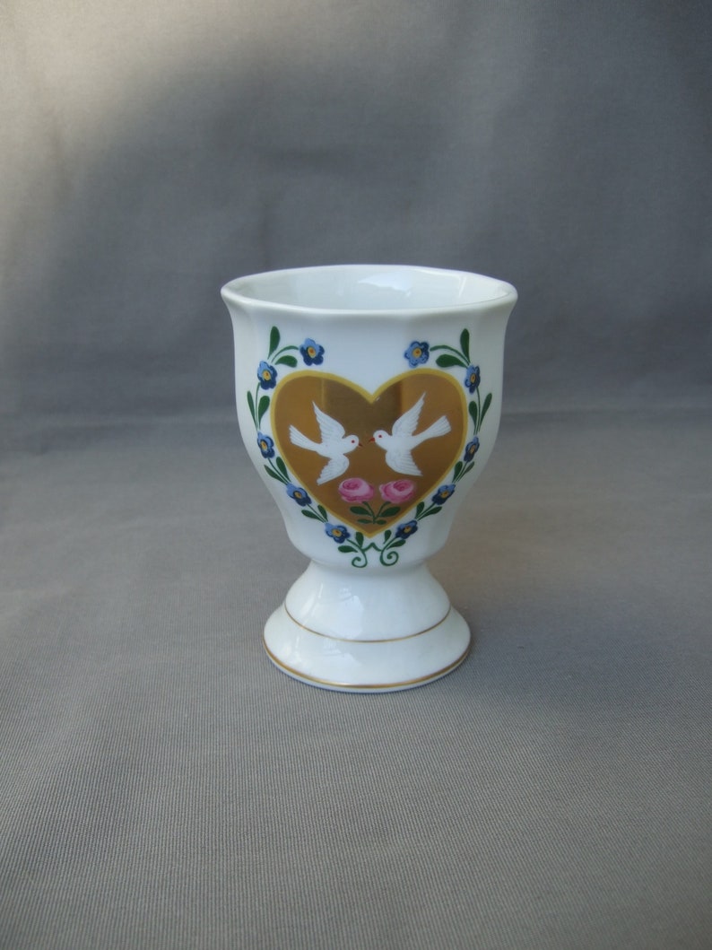 K&ouml;nnte beinhalten: Wei&szlig;e Keramiktasse mit einem goldenen Herzdesign mit zwei wei&szlig;en Tauben und rosa Rosen. Die Tasse hat einen goldenen Rand und ein blaues und gr&uuml;nes Blumenmuster.