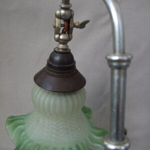 Op de afbeelding: Een bureaulamp met een gebogen zilverkleurige metalen arm en een matglazen kap met een lichtgroene, geplooide rand. De kap heeft een gestructureerd ribbelpatroon en is aan de metalen arm bevestigd met een donkerbruine fitting.