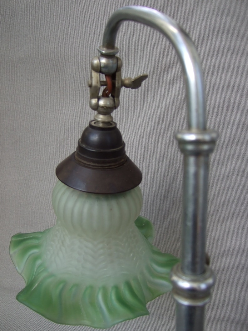 Op de afbeelding: Een bureaulamp met een gebogen zilverkleurige metalen arm en een matglazen kap met een lichtgroene, geplooide rand. De kap heeft een gestructureerd ribbelpatroon en is aan de metalen arm bevestigd met een donkerbruine fitting.