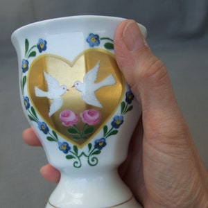 K&ouml;nnte beinhalten: Wei&szlig;e Keramiktasse mit einem goldenen Herzdesign mit zwei wei&szlig;en Tauben und rosa Blumen. Die Tasse hat ein blaues und gr&uuml;nes Blumenmuster am Rand.