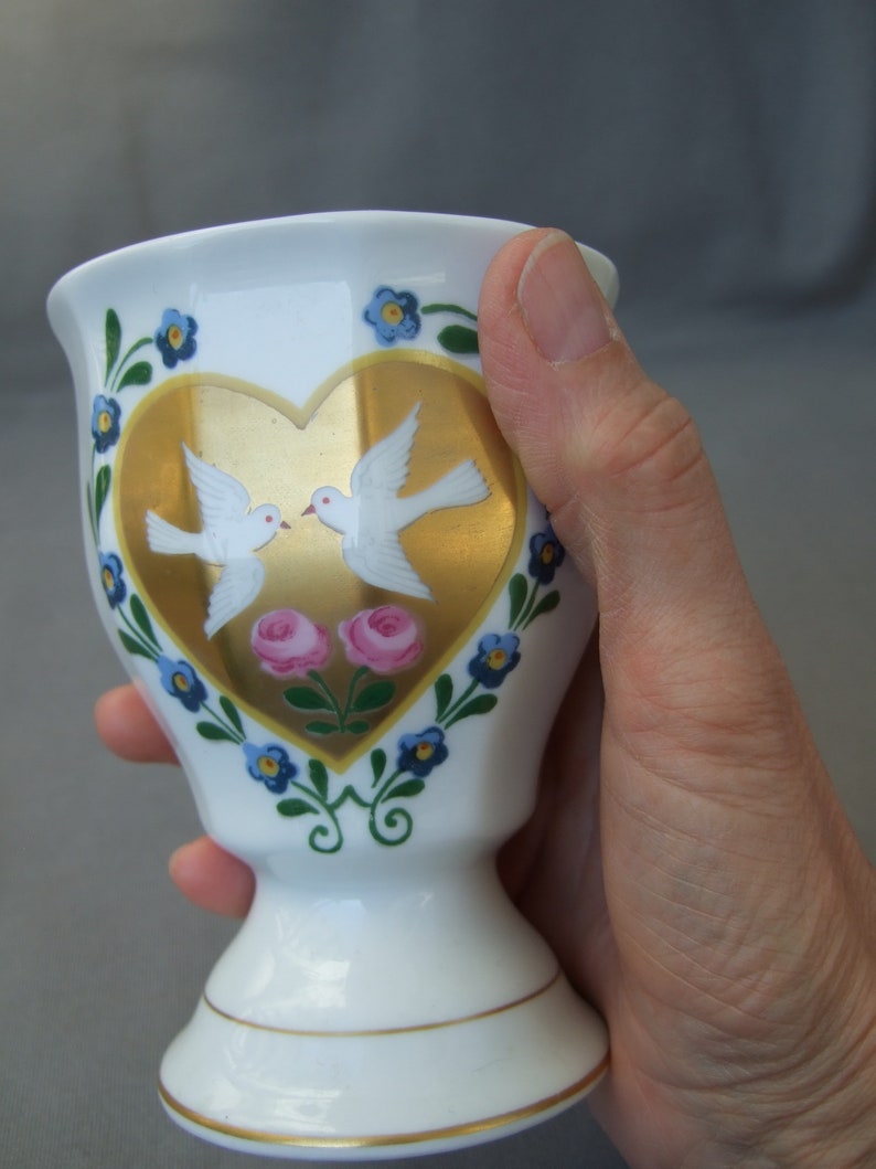 K&ouml;nnte beinhalten: Wei&szlig;e Keramiktasse mit einem goldenen Herzdesign mit zwei wei&szlig;en Tauben und rosa Blumen. Die Tasse hat ein blaues und gr&uuml;nes Blumenmuster am Rand.