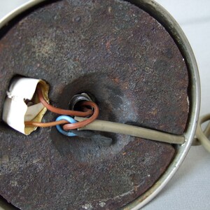 Op de afbeelding: De onderkant van een ronde lampvoet met een donker, getextureerd en roestig metalen gewicht. Een beige elektriciteitssnoer gaat de voet in, waarbij bruine en blauwe interne draden zichtbaar zijn bij een centrale opening en een klein wit papieren label.