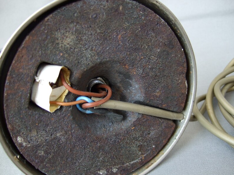 Op de afbeelding: De onderkant van een ronde lampvoet met een donker, getextureerd en roestig metalen gewicht. Een beige elektriciteitssnoer gaat de voet in, waarbij bruine en blauwe interne draden zichtbaar zijn bij een centrale opening en een klein wit papieren label.