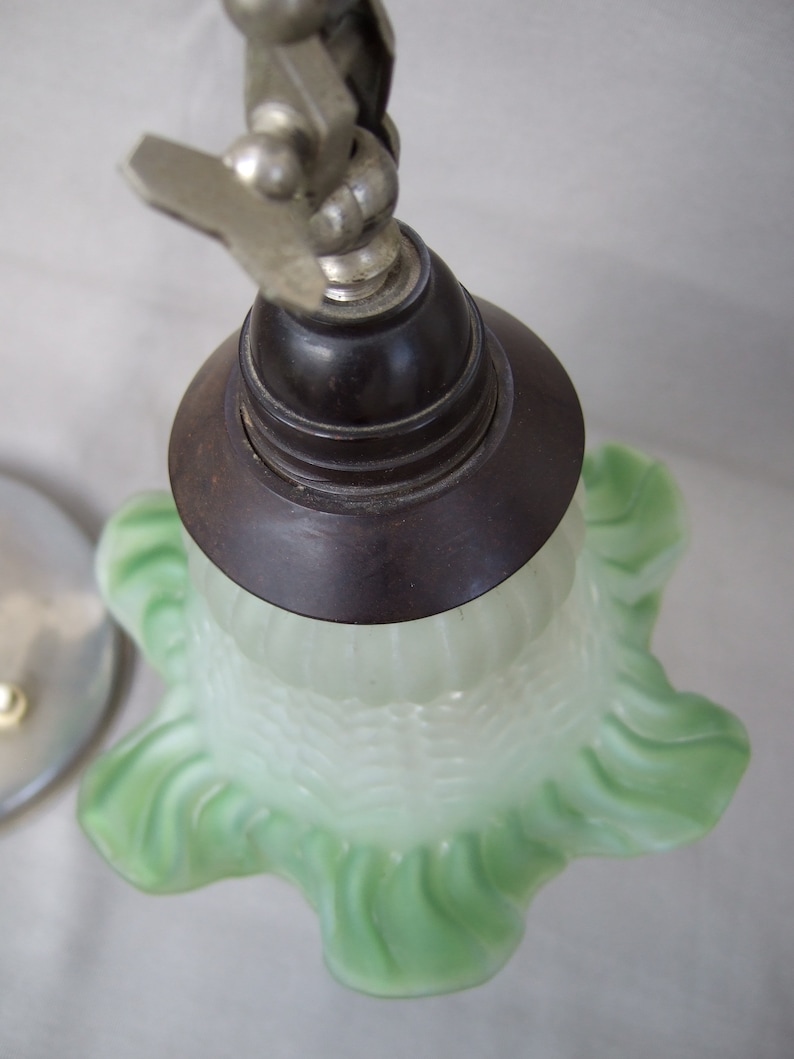 Op de afbeelding: Een lampenkap van matglas met een lichtgroene, gegolfde rand en een geribbelde textuur. De kap is bevestigd aan een donkerbruine ronde fitting, die verbonden is met een metalen scharnierarm met een zichtbare vleugelmoer aan de bovenzijde.