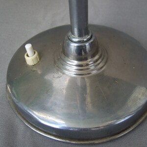 Op de afbeelding: Een ronde, gepolijste chromen lampvoet met een glad, reflecterend oppervlak. Een kleine, witte cilindrische drukknopschakelaar is bovenop de voet gemonteerd, links van de centrale verticale chromen lampstang geplaatst.