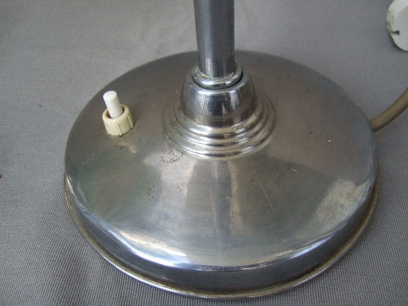 Op de afbeelding: Een ronde, gepolijste chromen lampvoet met een glad, reflecterend oppervlak. Een kleine, witte cilindrische drukknopschakelaar is bovenop de voet gemonteerd, links van de centrale verticale chromen lampstang geplaatst.