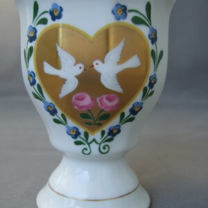 K&ouml;nnte beinhalten: Ein wei&szlig;er Porzellan-Eierbecher mit einem goldenen Herzdesign mit zwei wei&szlig;en Tauben und rosa Rosen. Das Herz ist von einem blauen und gr&uuml;nen floralen Rand umgeben.