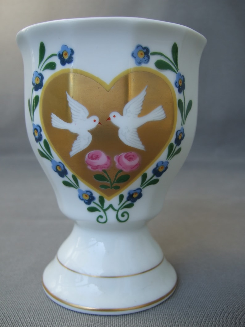 K&ouml;nnte beinhalten: Ein wei&szlig;er Porzellan-Eierbecher mit einem goldenen Herzdesign mit zwei wei&szlig;en Tauben und rosa Rosen. Das Herz ist von einem blauen und gr&uuml;nen floralen Rand umgeben.