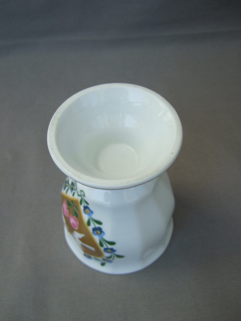 K&ouml;nnte beinhalten: Ein wei&szlig;er Keramik-Eierbecher mit floralem Design. Das Design zeigt rosa und blaue Blumen um einen goldenen Buchstaben "A".