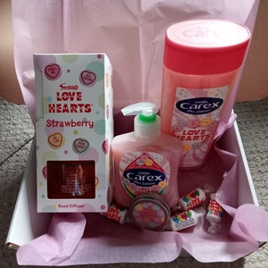 May include: A gift box filled with pink and white tissue paper. The box contains a strawberry scented reed diffuser, a bottle of Carex Fun Edition hand wash, a small tin of Love Hearts sweets, and several Love Hearts candies.